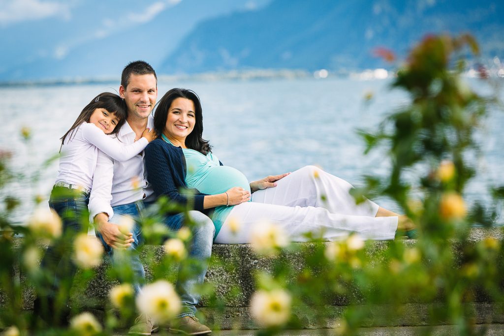 Family of three pose for a maternity photo on the Vevey lakeside framed by flowers